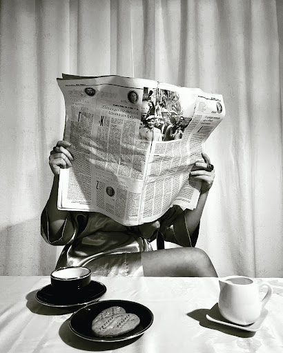 A black and white image of a woman sitting at a breakfast table reading a newspaper